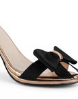 Vista a detalle de moño al frente de zapatilla negra de diseño descubierto para mujer con pulsera y tacón 10cm